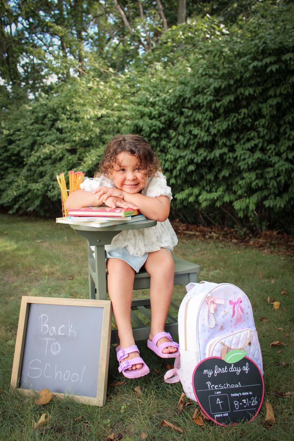 Preschool-aged child smiling beside school books, pencils, a backpack, and first-day signs